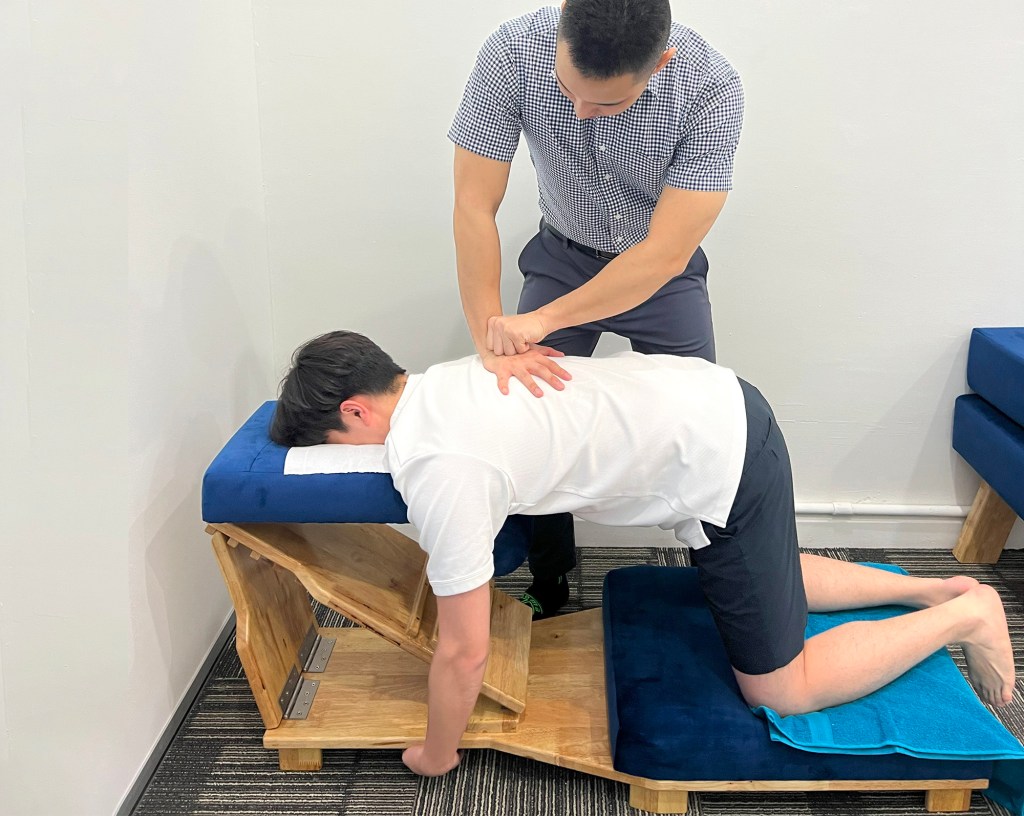 Photograph of Dr Kit adjusting a patient's back. The patient is lying face down on a chiropractic device, and Dr Kit has his palm on his upper middle back.